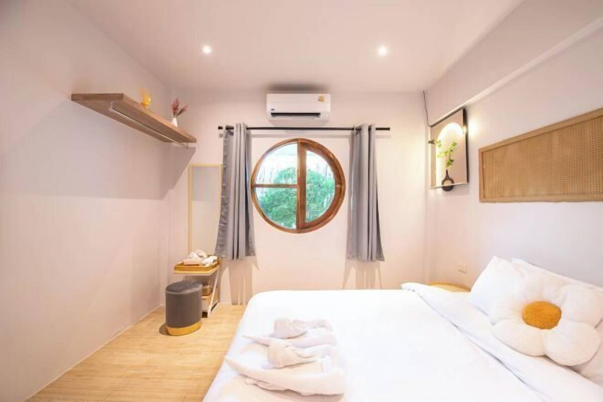 Une chambre d’hôtel avec un lit, une fenêtre ronde, une petite table avec une lampe et une poubelle.