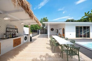 A modern outdoor kitchen with a dining area, a pool, and a thatched roof structure.