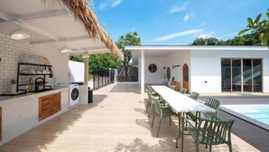 A modern outdoor kitchen area with a dining table and chairs, a pool, and a thatched roof structure. - De Beach pool villa. New Villa 3 min walk from the beach 5 rooms (Pattaya)