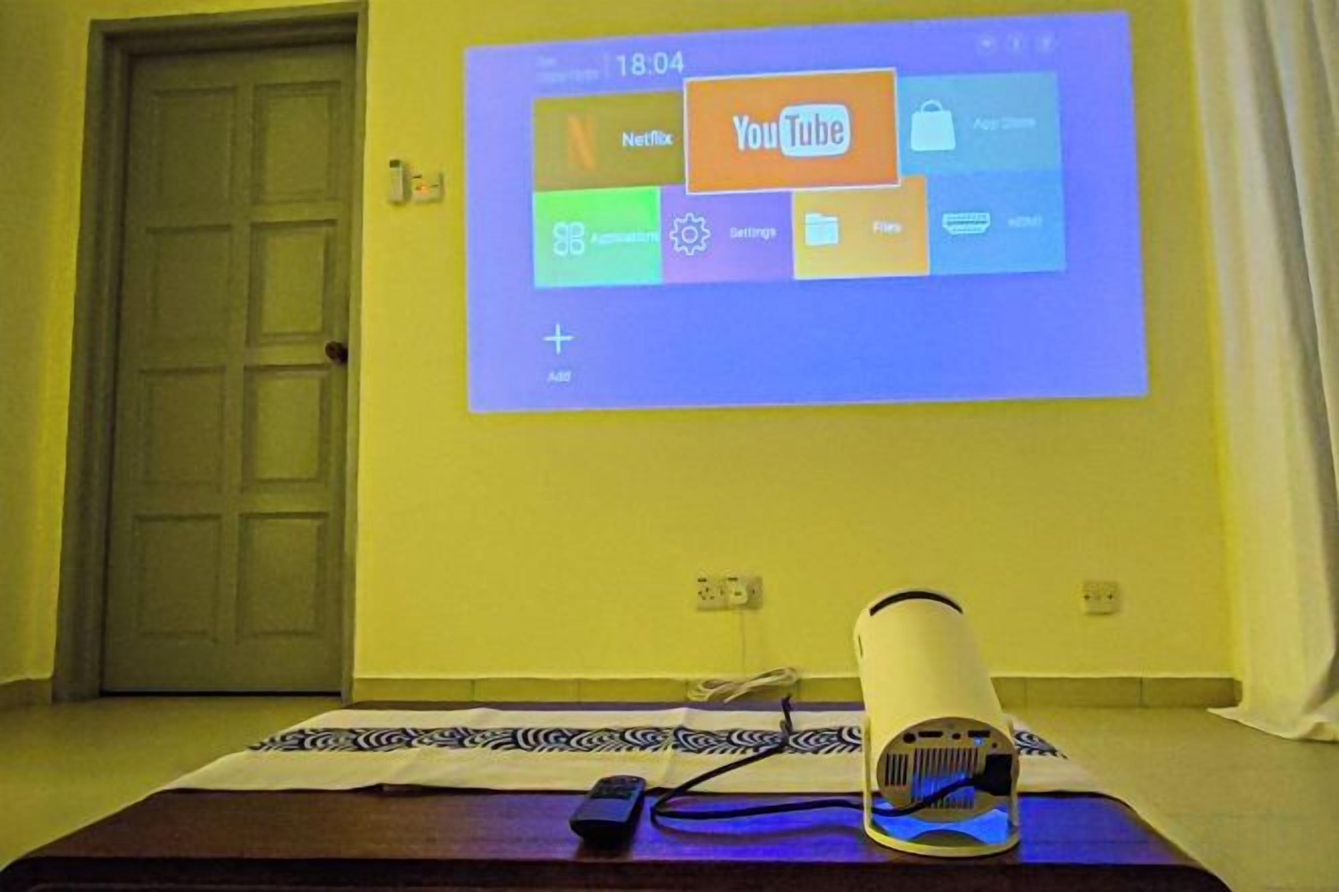 A projection screen displaying a YouTube interface on a wall, a remote control, and a speaker on a table.