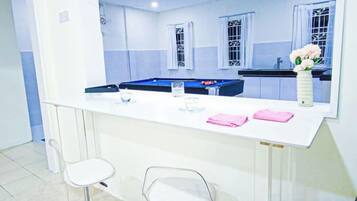 Habitación con barra blanca, dos taburetes, mesa de billar y ventana con vistas.