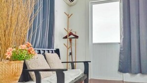 Una habitación con cama, perchero de madera, planta en maceta y ventana con cortinas.