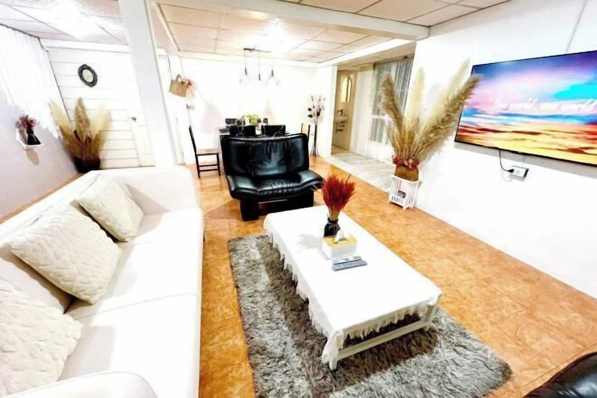 A modern living room with a white sofa, a black leather chair, a white coffee table, and a large TV on the wall.