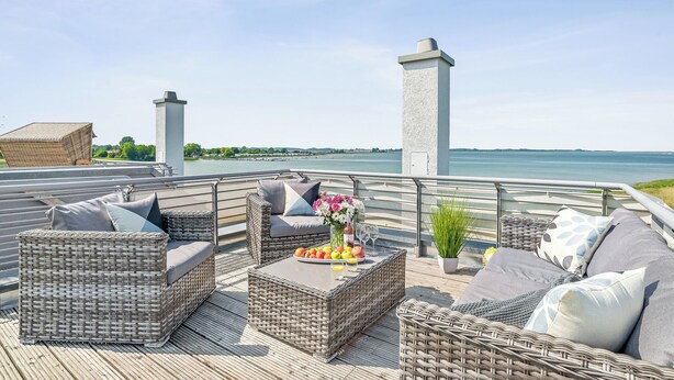 A deck with wicker furniture, a table with fruit and flowers, and a view of the water. - Vacation home Bellevue directly on the Schlei with fireplace, sauna and whirlpool (Kappeln)