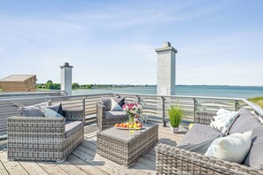 A deck with wicker furniture, a table with fruit and flowers, and a view of the water.