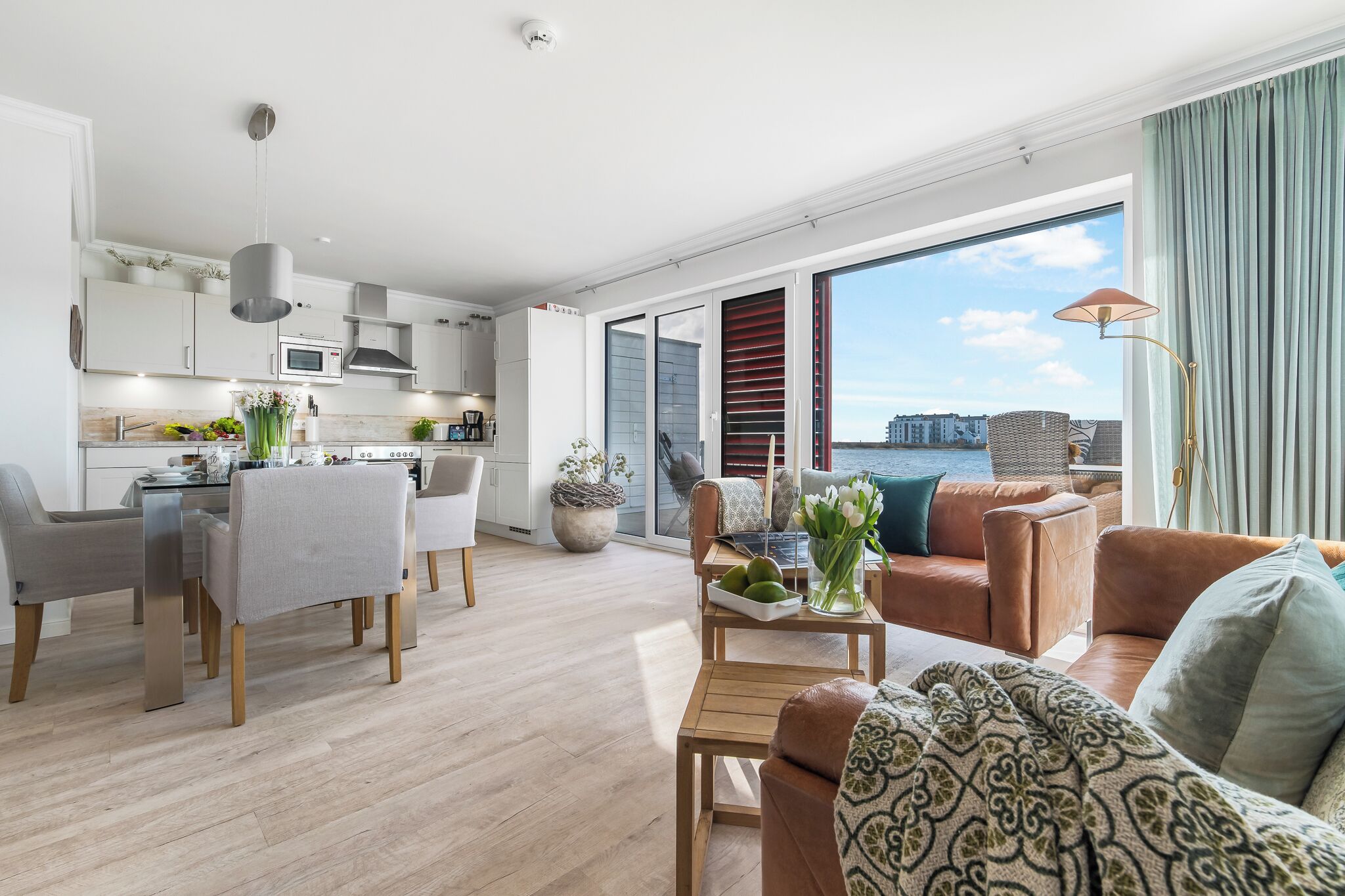 A modern living room with a sofa, coffee table, and dining area. A large window offers a view of the water and cityscape.
