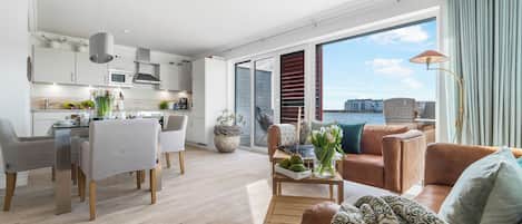 A modern living room with a sofa, coffee table, and dining area. A large window offers a view of the water and cityscape.