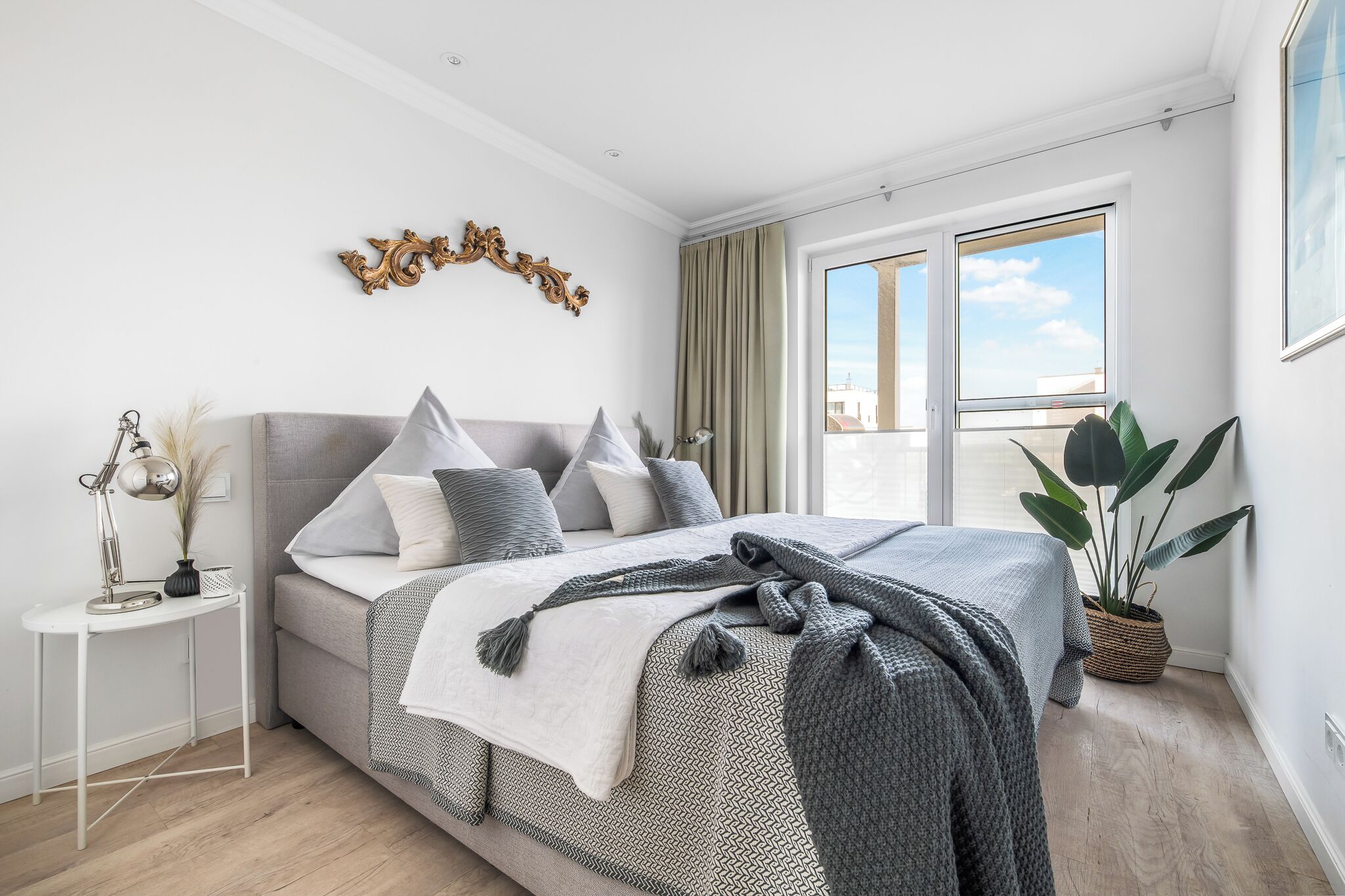 Quarto moderno com uma cama grande, uma mesinha de cabeceira, um objeto decorativo na parede e um vaso com planta.
