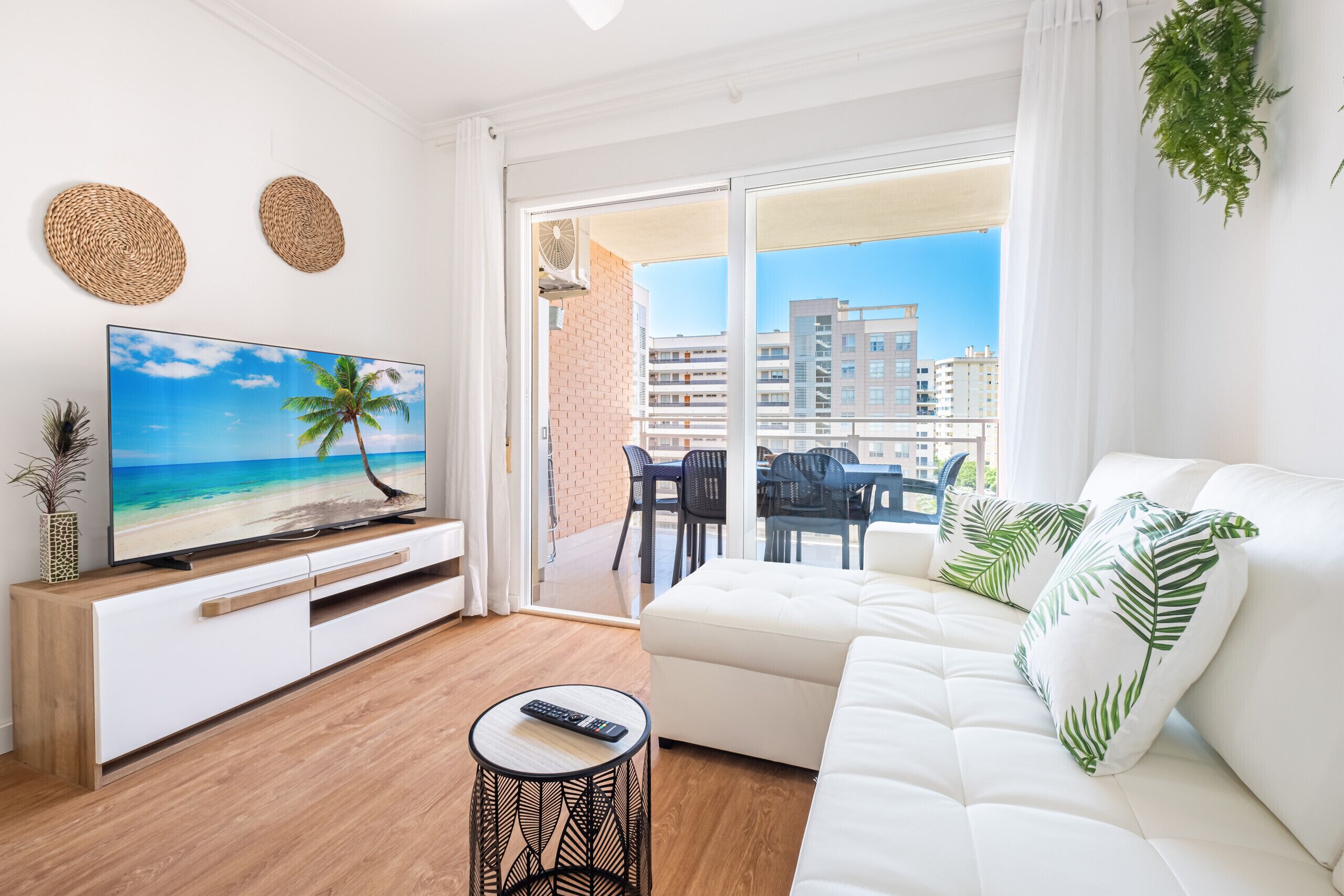 Un salon moderne doté d’un téléviseur à écran plat, d’un canapé blanc et d’un balcon offrant une vue sur les immeubles.