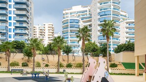 Un parque infantil con columpios y toboganes, rodeado de palmeras y edificios modernos al fondo.