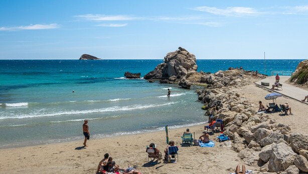 Una playa con agua azul cristalina, afloramientos rocosos y gente descansando sobre la arena.