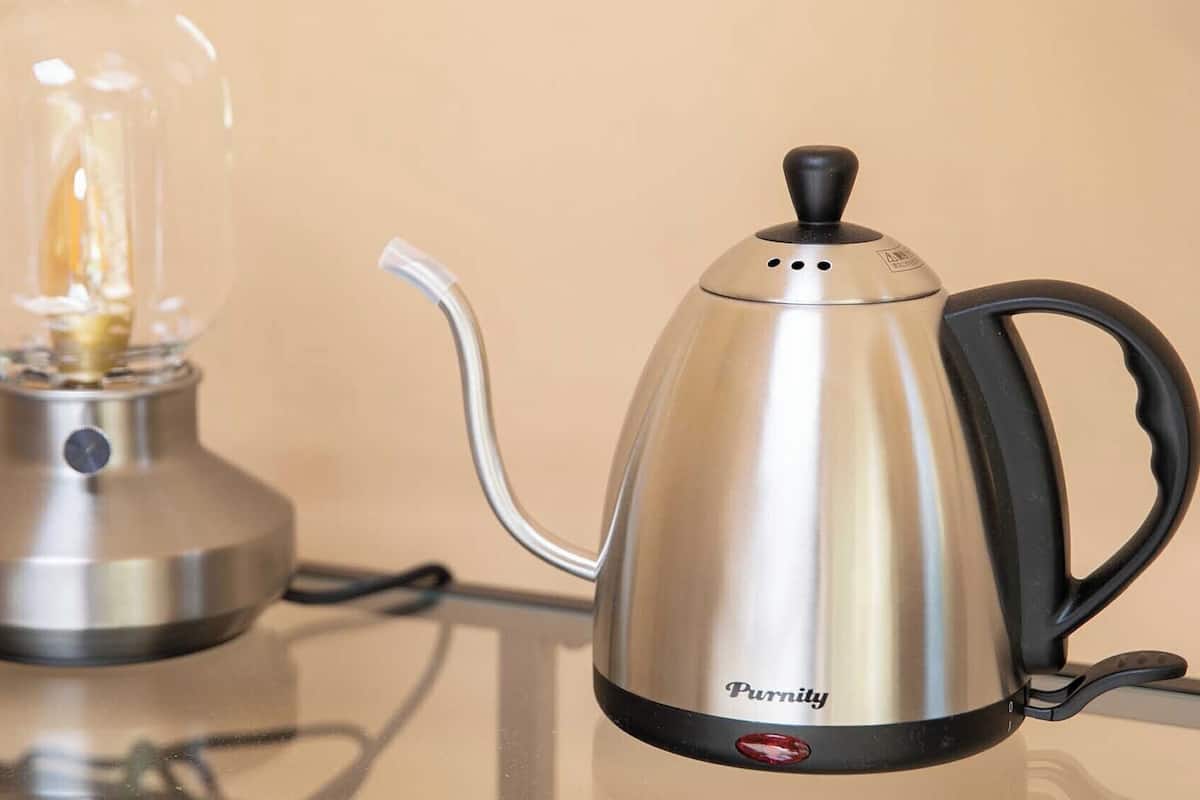 A stainless steel coffee pot with a black handle and a glass bulb light on a reflective surface.