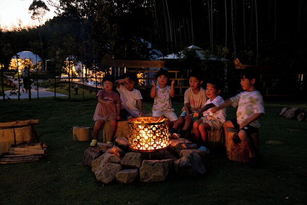 A group of children sitting around a campfire in a grassy outdoor area at dusk.