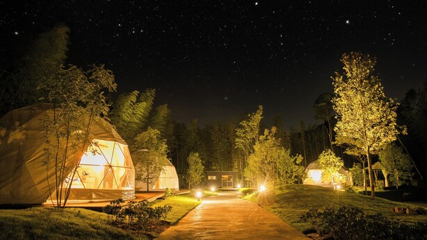 A nighttime scene with several dome-shaped tents illuminated by artificial lighting, set in a forested area with a clear path leading to a central building.