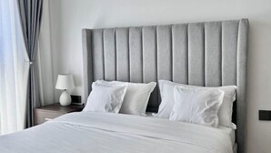 A bed with a grey upholstered headboard, white bedding, and a bedside lamp on a wooden nightstand. - Modern 1Br, Queen, Gym, Pool,Central in Kileleshwa (Nairobi)