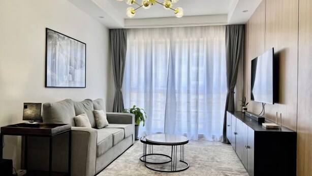 A modern living room with a sofa, a coffee table, a TV mounted on the wall, and a large window with curtains. - Modern 1Br, Queen, Gym, Pool,Central in Kileleshwa (Nairobi)