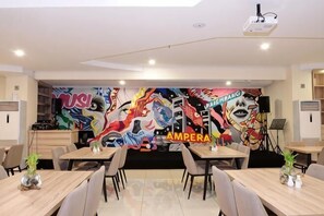 Un comedor con mesas y sillas, una pantalla para proyector y un mural de colores en la pared.
