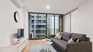 A modern living room with a grey sofa, a small white table with a plant, a TV on a white cabinet, and a large window with a view of buildings. - Charming 2BR Apartment with Free Parking (Melbourne)