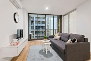 Un salon moderne avec un canapé gris, une petite table blanche ornée d’une plante, un téléviseur posé sur un meuble blanc et une grande fenêtre donnant sur des immeubles.