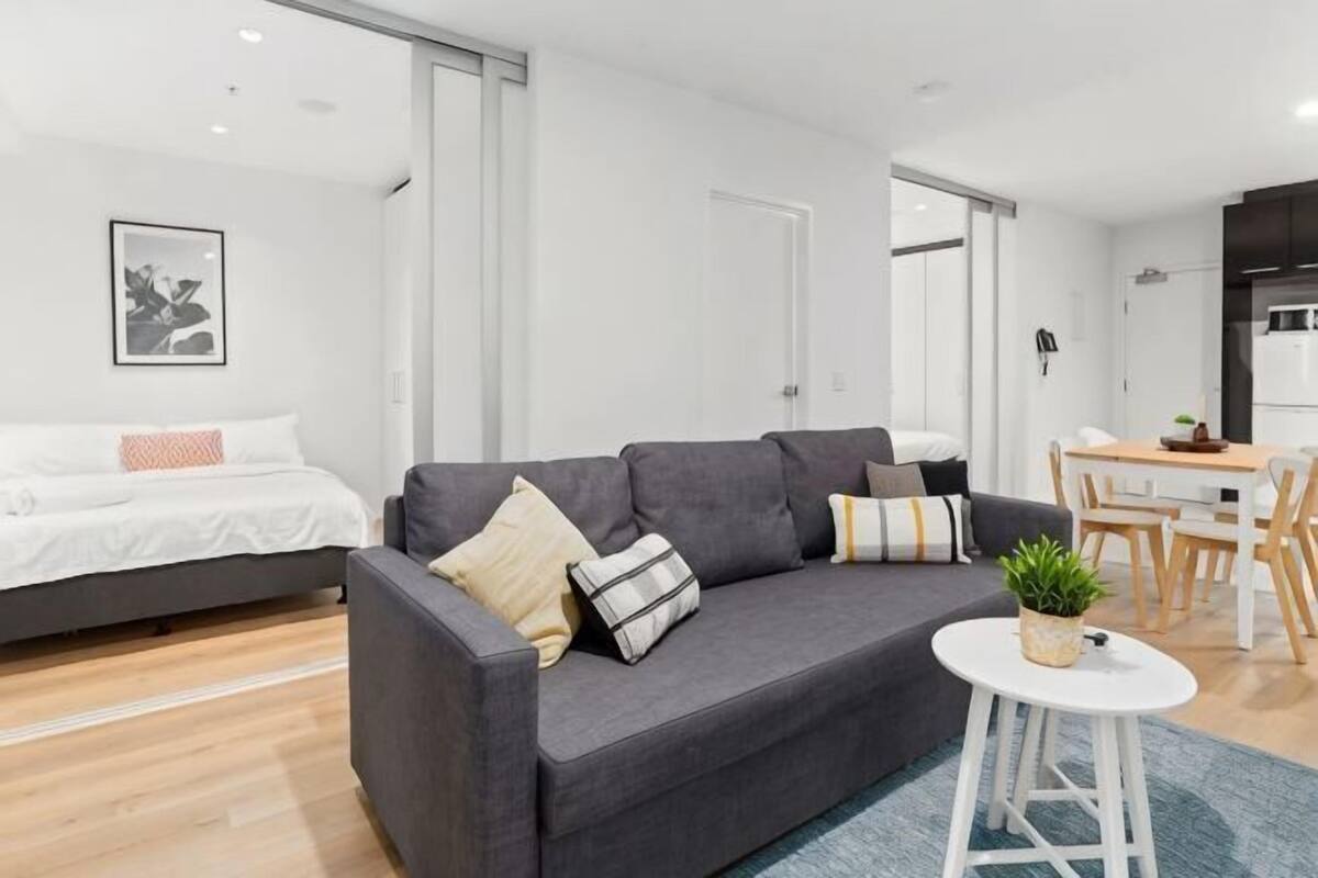 Een moderne woonkamer met een bank, een klein rond tafeltje met een plant, een bed met een witte hoofdbord en een eethoek met een houten tafel en stoelen.