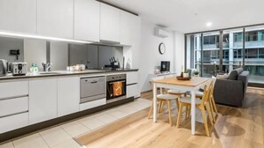 A modern kitchen with white cabinets, a dining area with a wooden table and chairs, and a large window with a view of the city. - Charming 2BR Apartment with Free Parking (Melbourne)