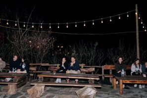 Un área de estar al aire libre con bancos y mesas de madera, luces colgantes y un cielo nocturno oscuro.