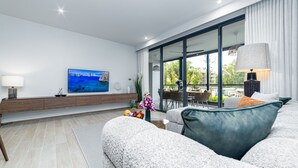 A modern living room with a large flat-screen TV, a sofa, a coffee table, and a dining area with a view of the outdoors. - 2BR Beachside 200 m to BangTao beach (Choeng Thale)