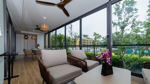A modern room with wicker furniture, a ceiling fan, large windows, and a view of greenery and buildings. - 2BR Beachside 200 m to BangTao beach (Choeng Thale)