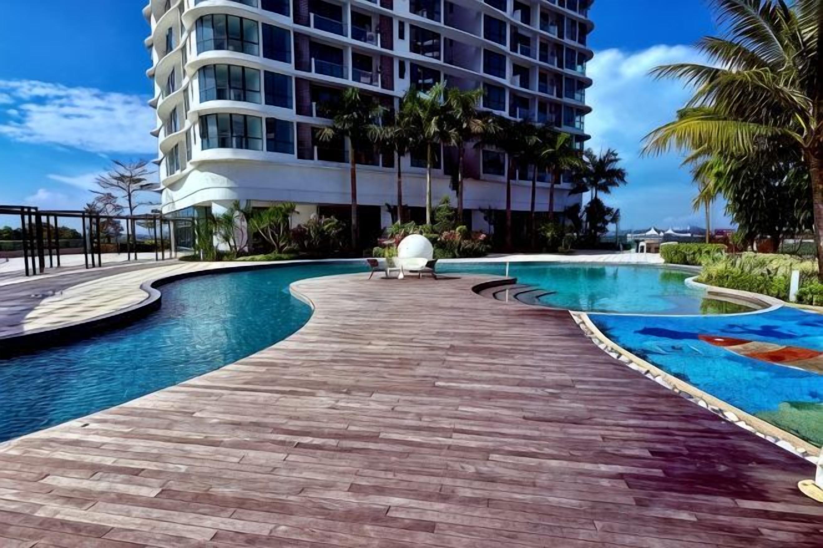 A swimming pool with a wooden deck, palm trees, and a modern hotel building in the background.