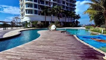 A swimming pool with a wooden deck, palm trees, and a modern hotel building in the background.