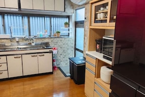 A compact kitchen with a sink, countertop, and cabinets. There is a window with blinds, a refrigerator, and a small trash bin.