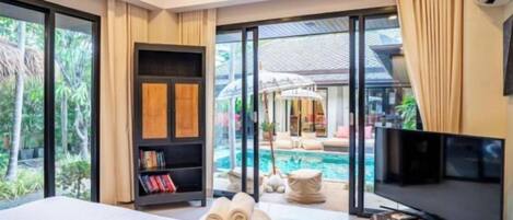 A hotel room with a bed, a television, a bookshelf, a pool view, and a large window.