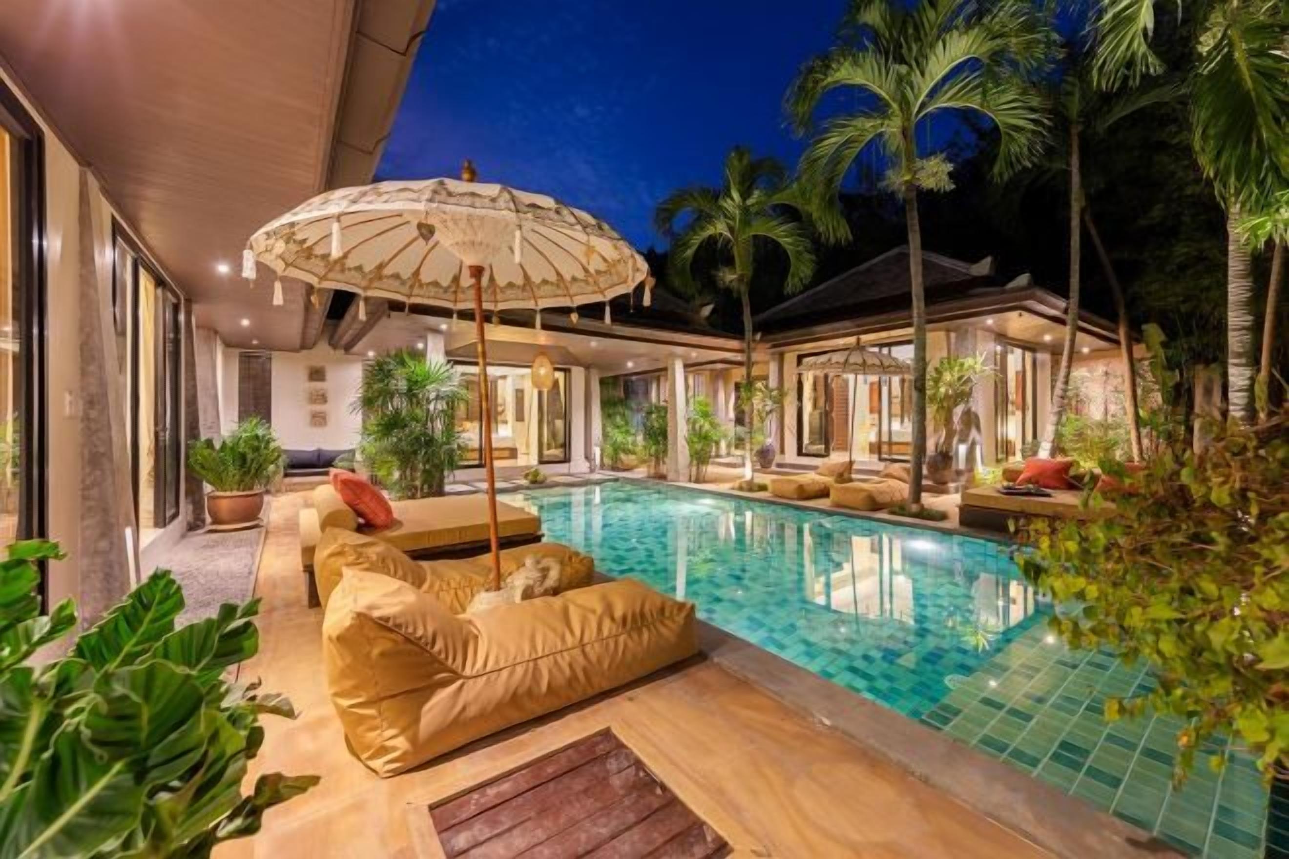 A pool area with a lounge chair and umbrella, surrounded by palm trees and a building with large windows.