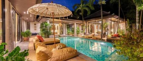 A pool area with a lounge chair and umbrella, surrounded by palm trees and a building with large windows.