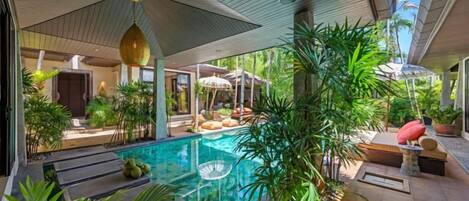 A modern outdoor pool area with a glass barrier, surrounded by tropical plants and a seating area with umbrellas.