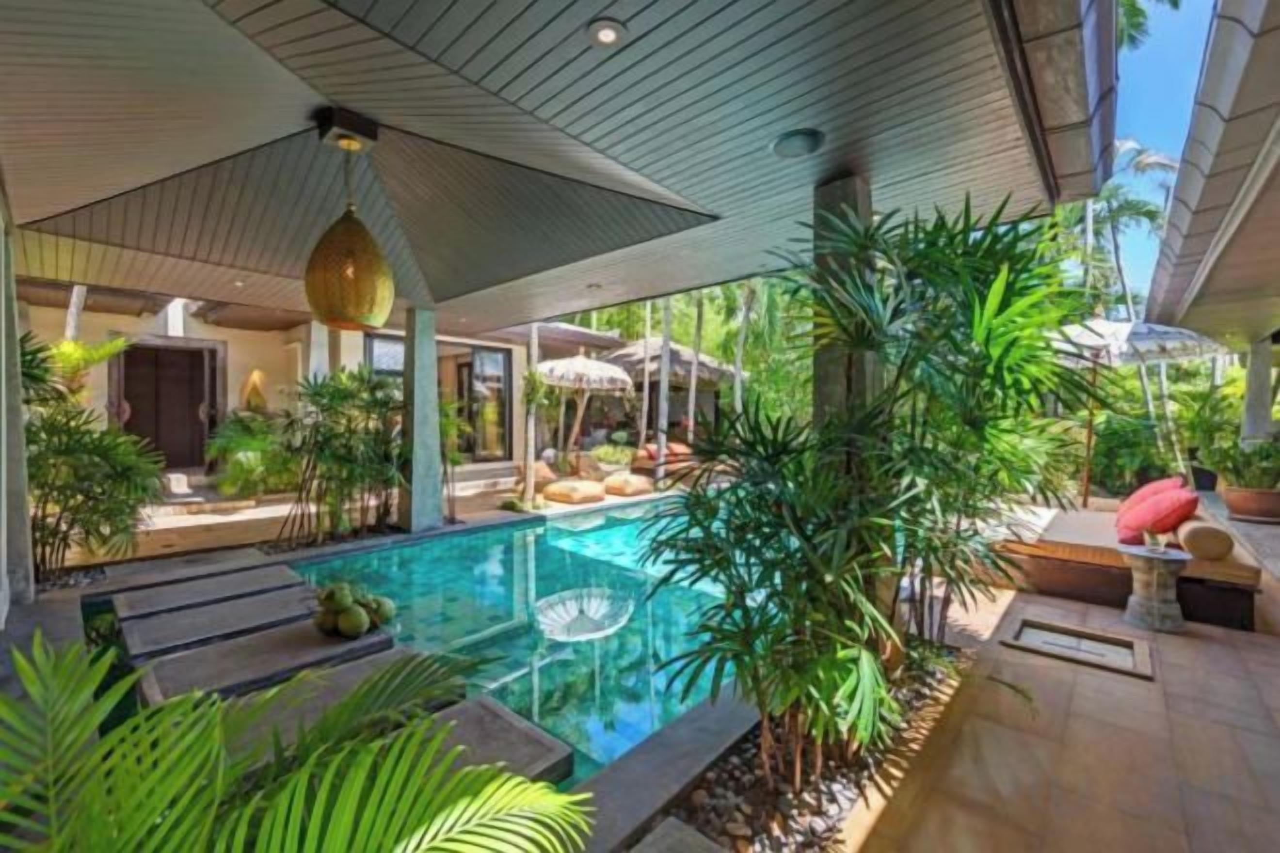 Un área de piscina moderna con una piscina cristalina, rodeada de plantas tropicales y un espacio de descanso cubierto.
