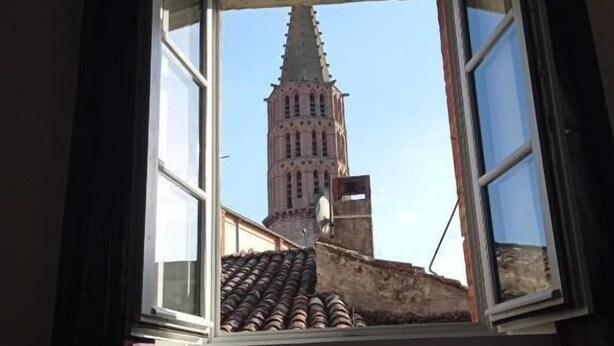 Una habitació amb una finestra oberta que dona a un edifici històric amb teulada apuntada.