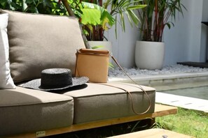 A beige sofa with a woven bag and a hat on it, positioned near a small pool with potted plants in the background. - Tatkala Villa (Bali)