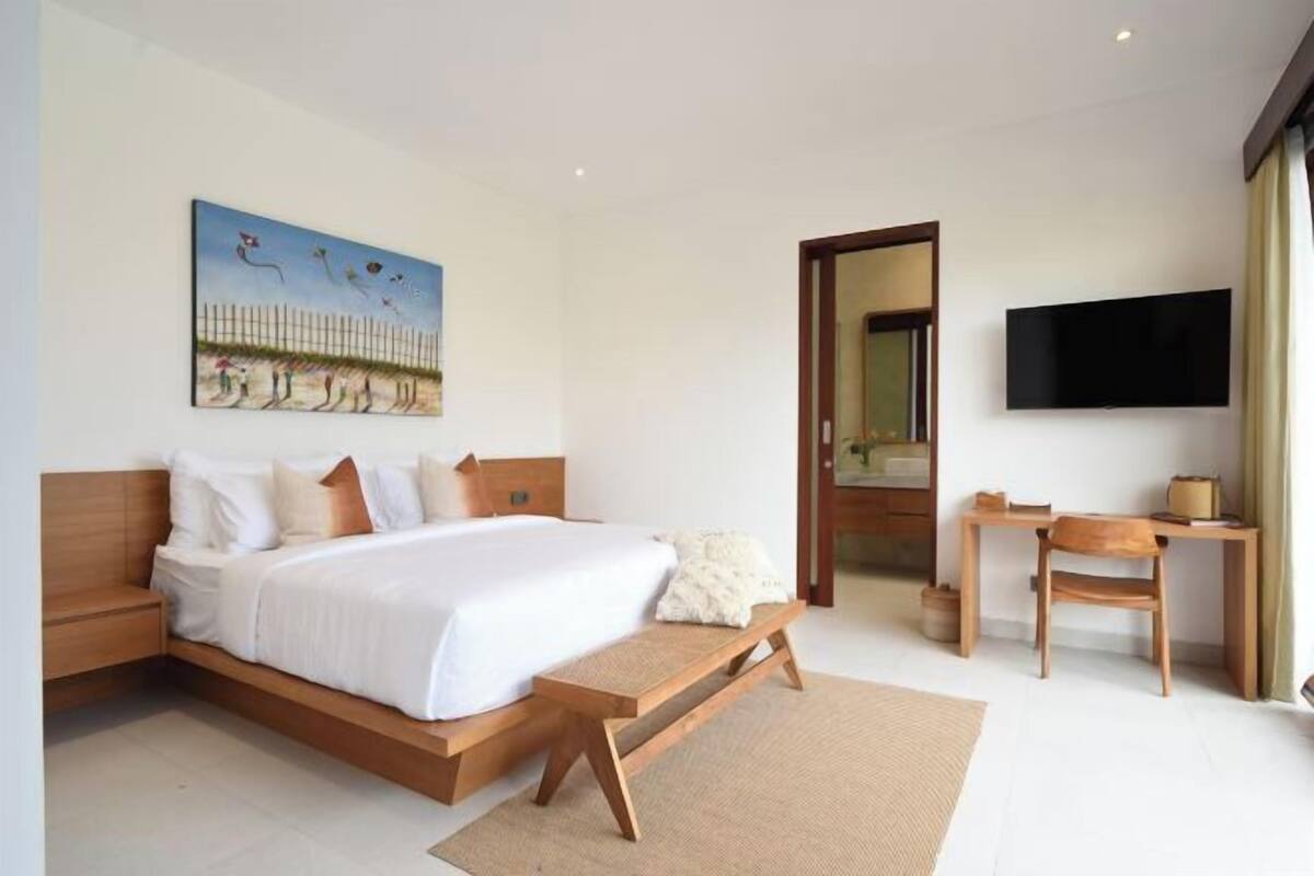 A bedroom with a bed, a wooden bench, a TV, and a painting on the wall.