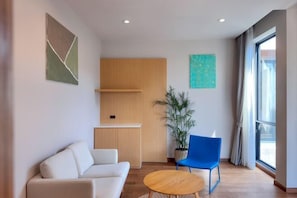 Una habitación moderna con sofá, una silla azul, una mesita, un armario de madera, una planta en maceta y una ventana con cortinas.