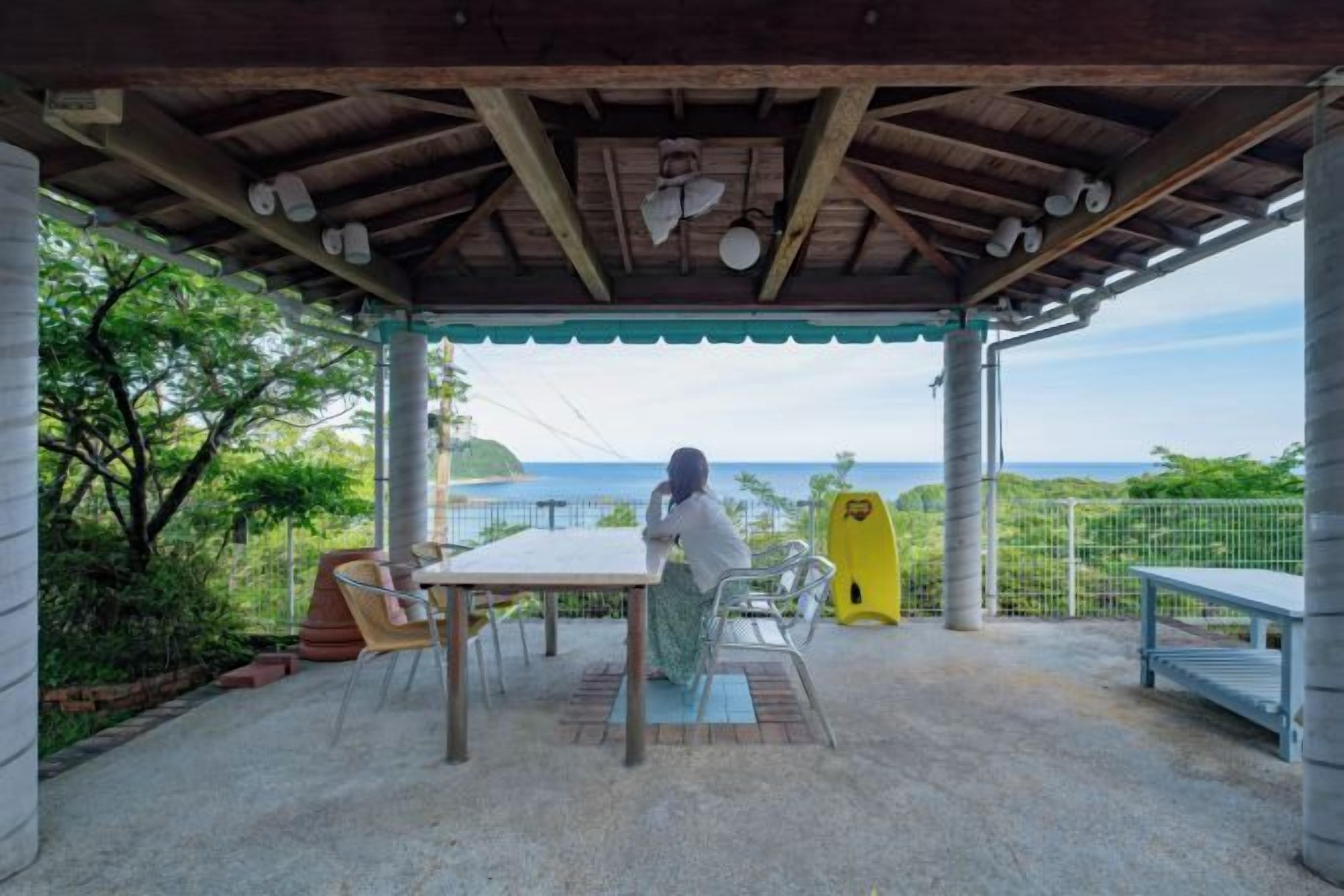 An outdoor patio with a table and chairs, a person sitting, and a view of the sea.