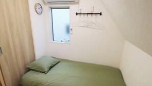 Een kleine kamer met een enkel bed, een groene plaid, een raam, een klok aan de muur en een kledingrek.