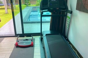 Une salle de sport à domicile équipée d’un tapis de course et d’un vélo d’appartement, d’un tapis de yoga, et offrant une vue sur une piscine grâce à de grandes fenêtres.