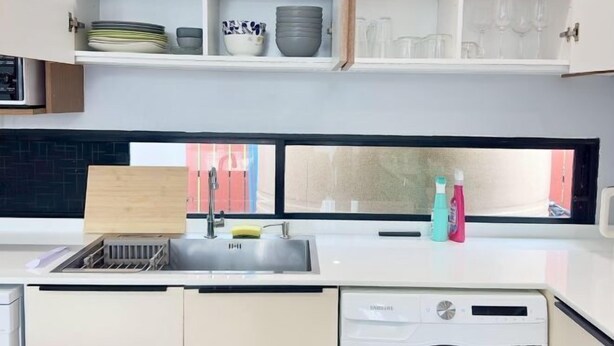 A modern kitchen with a built-in dishwasher, a sink with a dish rack, and a window with a view of a red building. - The White House Kamala Phuket (Phuket)