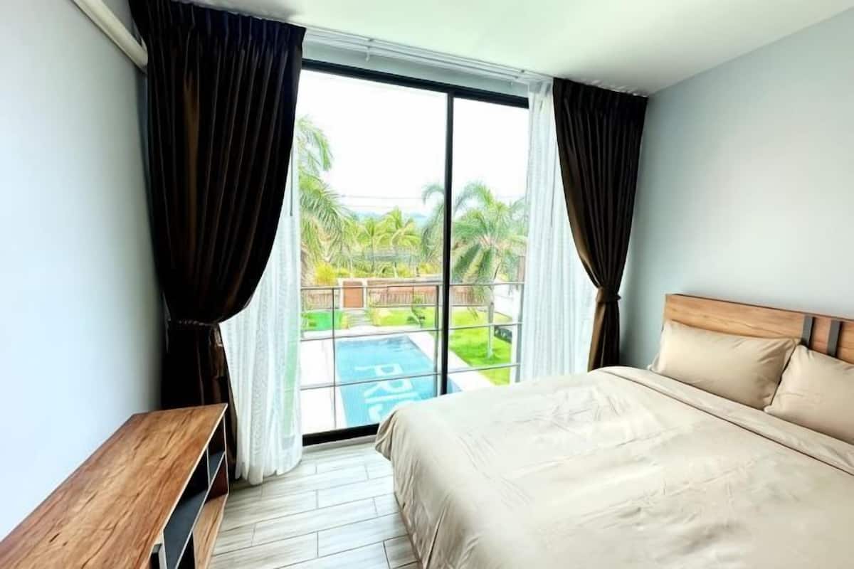Ein Schlafzimmer mit einem Bett, einer hölzernen Sitzbank, einem Fenster mit Vorhängen sowie Blick auf einen Pool und Grünflächen.