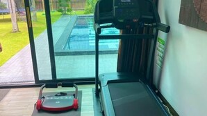 A treadmill and a stationary bike in a gym with large windows overlooking a pool. - The White House Kamala Phuket (Phuket)