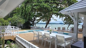 An outdoor pool area with white chairs and tables, surrounded by lush greenery and overlooking a body of water. - Seaside Bliss Villa Rawai (Phuket)
