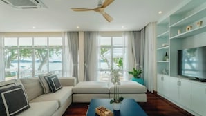 A modern living room with a sofa, a coffee table, and a TV. There are large windows with curtains, a ceiling fan, and a view of the outdoors. - Seaside Bliss Villa Rawai (Phuket)
