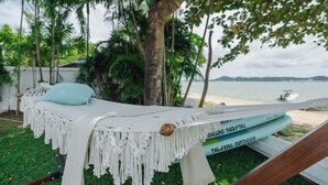 A hammock with macrame fringe, a light blue cushion, and a white cover, set against a backdrop of a beach and palm trees. - Seaside Bliss Villa Rawai (Phuket)