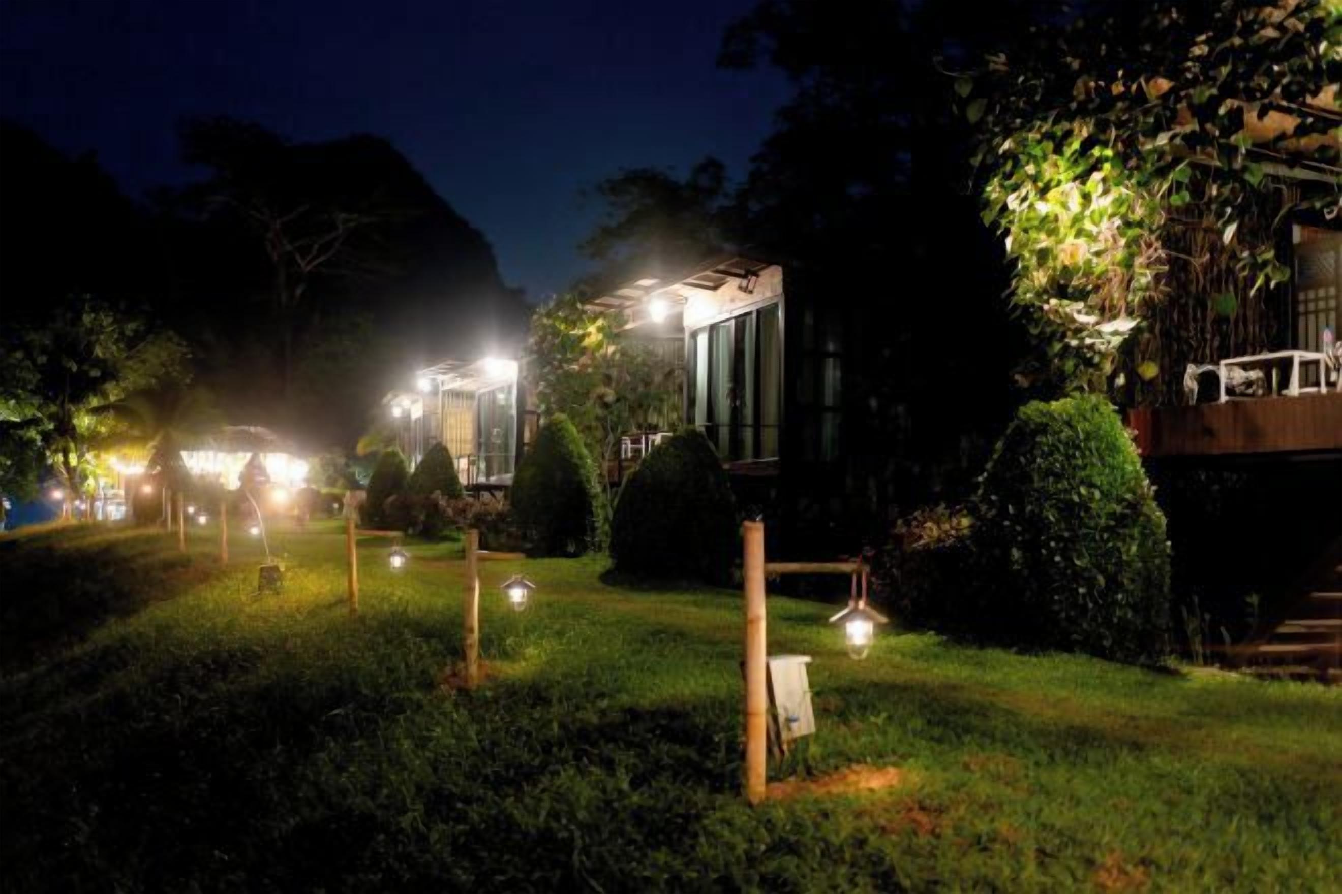 A well-lit outdoor area with a pathway leading to a building, surrounded by greenery and illuminated by multiple light sources.